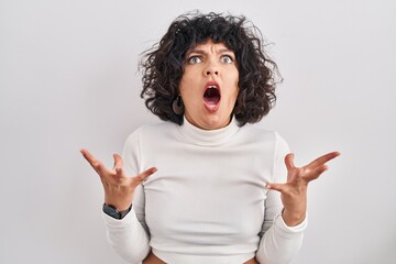 Hispanic woman with curly hair standing over isolated background crazy and mad shouting and yelling with aggressive expression and arms raised. frustration concept.