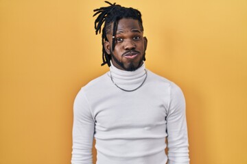 African man with dreadlocks wearing turtleneck sweater over yellow background puffing cheeks with...