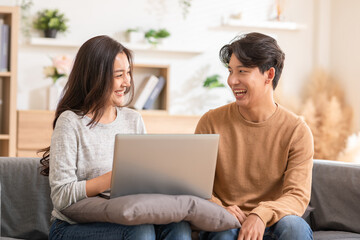 Asian young couple sitting on couch in living room enjoy with computer laptop spending time together. Happy couple surf social media at home both are smiling, laughing and enjoying moment on weekend.