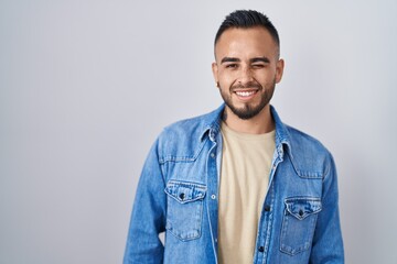 Fototapeta premium Young hispanic man standing over isolated background winking looking at the camera with sexy expression, cheerful and happy face.