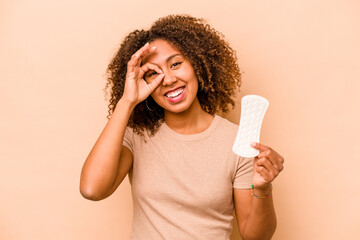 Young African American woman holding sanitary napkin isolated on beige background excited keeping ok gesture on eye.