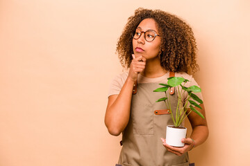 Young gardener African American woman holding plant isolated on beige background looking sideways...