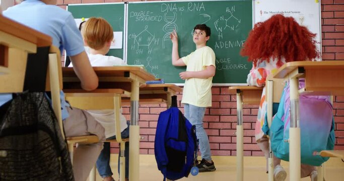 Caucasian smart teenager schoolboy answering at blackboard to woman teacher of biology and solving biological formulas. Class concept. Education for children. Pupils applauding in classroom.