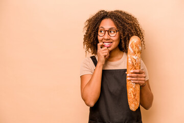 Young baker African woman holding a loaf of bread isolated on beige background relaxed thinking...