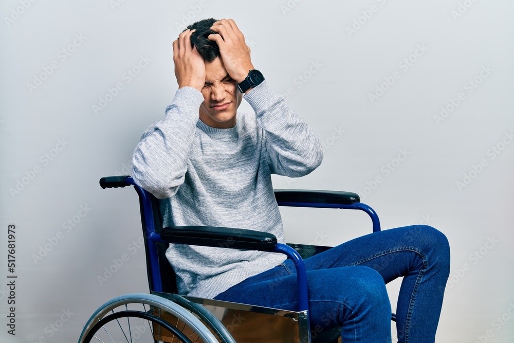 Sticker young hispanic man sitting on wheelchair suffering from headache desperate and stressed because pain