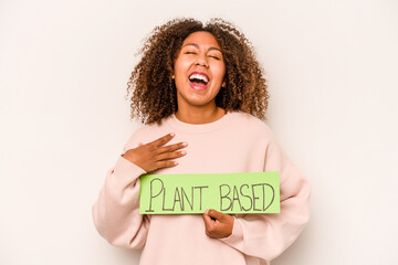 Young African American woman holding a plant based placard isolated on white background laughs out loudly keeping hand on chest.