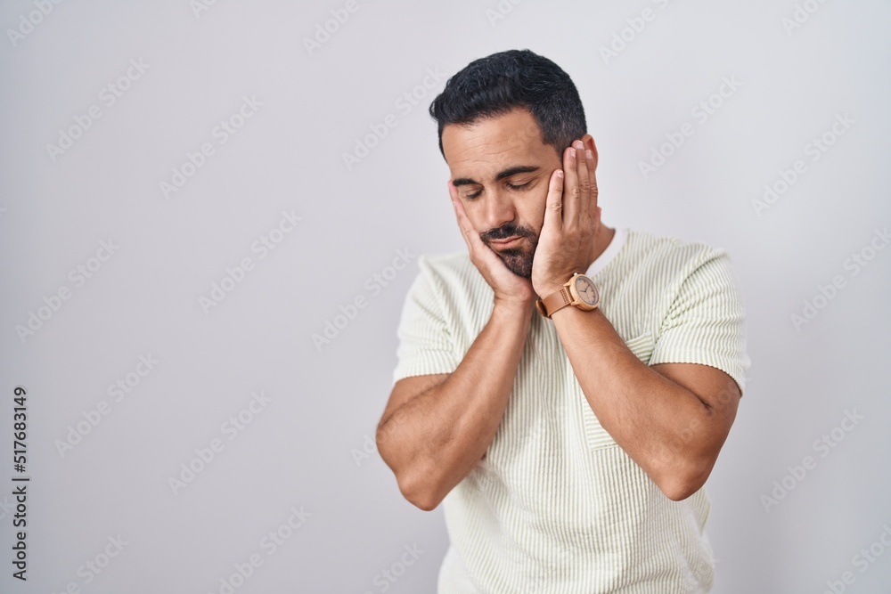 Wall mural Hispanic man with beard standing over isolated background tired hands covering face, depression and sadness, upset and irritated for problem