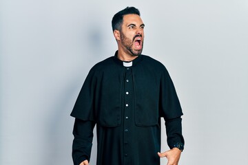 Handsome hispanic man with beard wearing catholic priest robe angry and mad screaming frustrated...