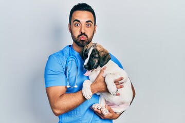 Handsome hispanic veterinary man with beard checking dog health puffing cheeks with funny face....