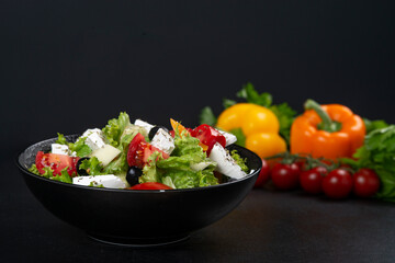 Healthy greek salad of fresh vegetables with tomatoes, lettuce, bell pepper, cucumbers, olives, feta cheese in plate on black stone background. Healthy food, vegetarian dieting, close up