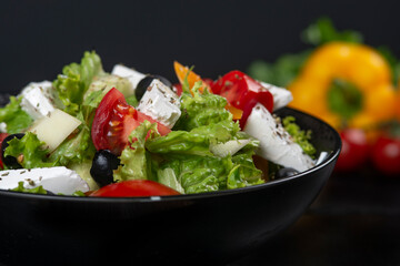 Healthy greek salad of fresh vegetables with tomatoes, lettuce, bell pepper, cucumbers, olives, feta cheese in plate on black stone background. Healthy food, vegetarian dieting, close up