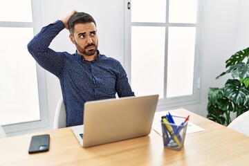 Young hispanic man with beard working at the office with laptop confuse and wondering about question. uncertain with doubt, thinking with hand on head. pensive concept.