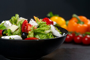 Healthy greek salad of fresh vegetables with tomatoes, lettuce, bell pepper, cucumbers, olives, feta cheese in plate on black stone background. Healthy food, vegetarian dieting, close up