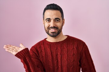 Fototapeta premium Young hispanic man with beard wearing casual sweater over pink background smiling cheerful presenting and pointing with palm of hand looking at the camera.