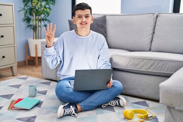 Non binary person studying using computer laptop sitting on the floor showing and pointing up with fingers number four while smiling confident and happy.