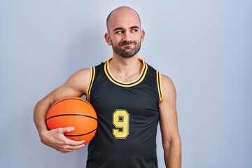 Young bald man with beard wearing basketball uniform holding ball smiling looking to the side and staring away thinking.