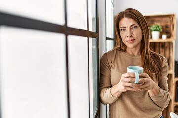 Middle age caucasian woman with serious expression drinking coffee standing at home.