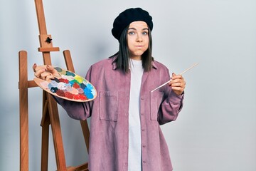 Young brunette woman standing by empty easel stand holding palette puffing cheeks with funny face. mouth inflated with air, catching air.