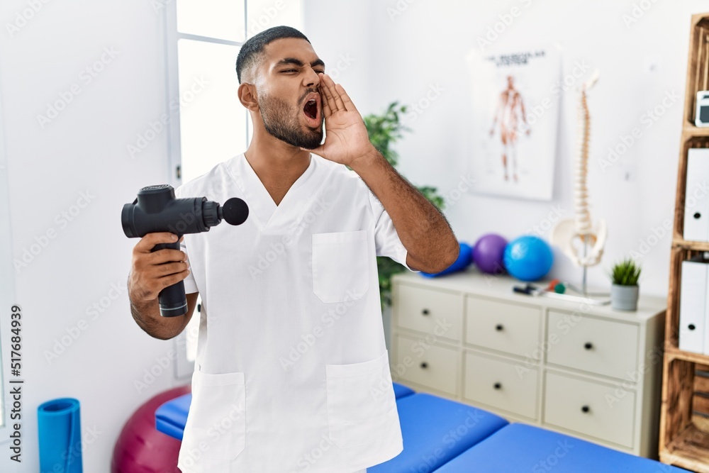 Poster young indian physiotherapist holding therapy massage gun at wellness center shouting and screaming l