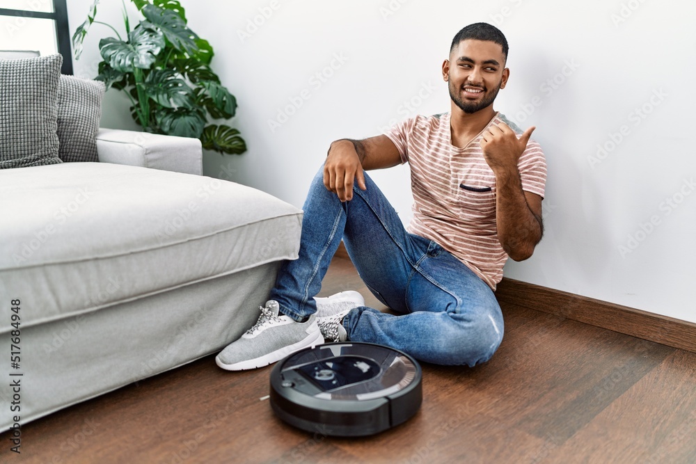 Poster young indian man sitting at home by vacuum robot smiling with happy face looking and pointing to the