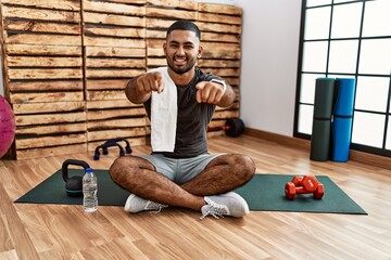 Young indian man sitting on training mat at the gym pointing to you and the camera with fingers,...