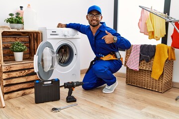 Young indian technician working on washing machine looking confident with smile on face, pointing...