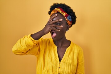 African young woman wearing african turban peeking in shock covering face and eyes with hand, looking through fingers with embarrassed expression.