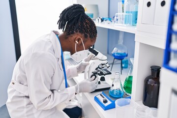 African american woman wearing scientist uniform and medical mask using microscope at laboratory