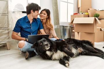 Young caucasian couple using laptop sitting on the floor with dog at new home.