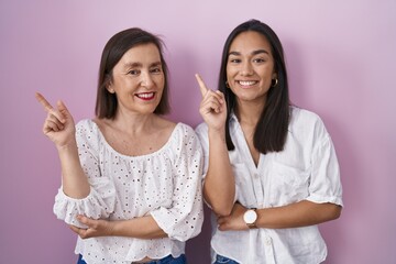 Hispanic mother and daughter together with a big smile on face, pointing with hand finger to the side looking at the camera.