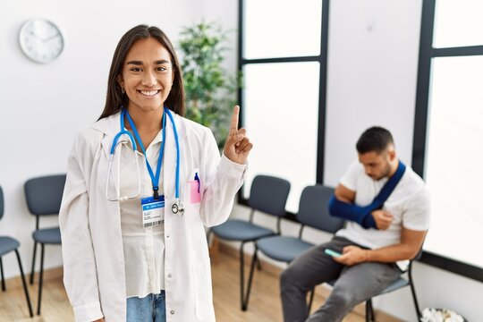 Young asian doctor woman at waiting room with a man with a broken arm showing and pointing up with finger number one while smiling confident and happy.