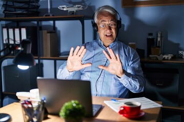 Fototapeta premium Hispanic senior man wearing call center agent headset at night afraid and terrified with fear expression stop gesture with hands, shouting in shock. panic concept.