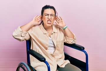 Young hispanic woman sitting on wheelchair trying to hear both hands on ear gesture, curious for gossip. hearing problem, deaf