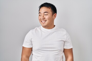 Young chinese man standing over white background looking away to side with smile on face, natural expression. laughing confident.