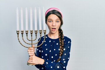 Young brunette girl holding menorah hanukkah jewish candle afraid and shocked with surprise and...