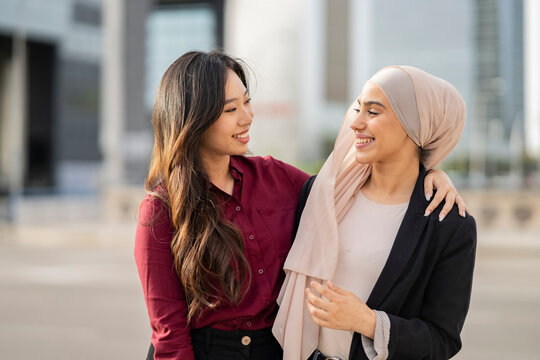 multiracial businesswomen friends hug outside, asian woman with muslim woman in buildings.
