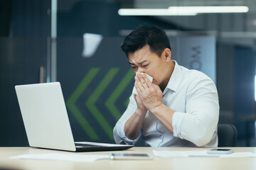 Sick man in office coughs and has runny nose, Asian businessman with cold works in office on laptop