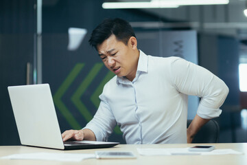 Asian businessman working in office, having severe back pain, overtired worker working with laptop.