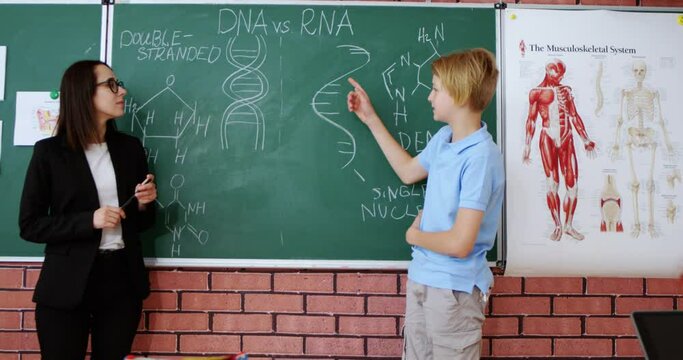 Caucasian clever teenage schoolboy answering at blackboard to female teacher of biology and solving biological formulas. Class concept. Education for children. Pupils applauding in classroom.