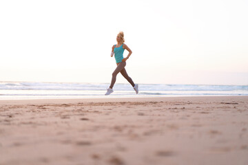 Active sporty woman run along ocean surf by water pool to keep fit and health. Sunset sand beach background with sun. Woman fitness, jogging workout and sport activity