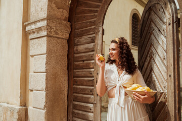 Happy smiling curly brunette woman wearing trendy summer white linen shirt, skirt, yellow headband, holding bowl with lemons, posing in sunny street of European city. Copy, empty space for text