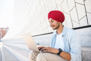 Side view at young modern Indian freelancer guy in national turban using laptop, hindu developer man in casual shirt typing sitting outdoors in cityscape, codding, working remotely on outsourcing - obrazy, fototapety, plakaty