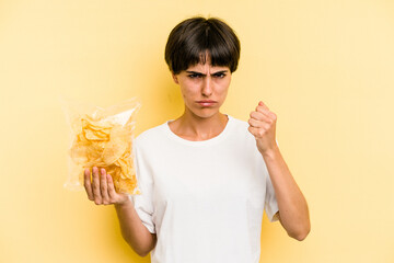 Young caucasian man holding crisps isolated on yellow background showing fist to camera, aggressive...