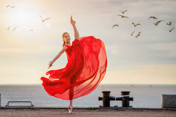 Dancing ballerina in a red flying skirt and leotard on the ocean embankment or on the sea beach...