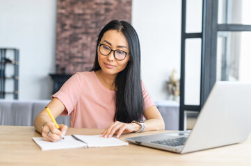 Mature asiatic woman receives a second education online, studies remotely, writes notes, looks...