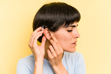 Young caucasian woman wearing a hearing aid isolated on yellow background