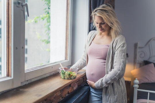 Caucasian pregnant woman with vegetable salad standing near the window, cowboy shot pregnancy and healthy food concept. High quality photo