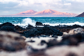 Wild rocky coastline of Fuerteventura and Lanzarote, Canary Islands, Spain. Playa Blanca on Lanzarote with volcano mountain iluminated in sunset seen from Corralejo - obrazy, fototapety, plakaty