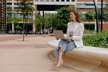 Fototapeta premium Adorable lovely girl with wonderful smile wearing white jacket and jeans sitting outdoor with laptop and working remote on background of modern building 