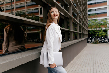 Smiling confident woman with long hair wraith white jacket is holding laptop and posing on background of modern business district in warm sunny day - obrazy, fototapety, plakaty